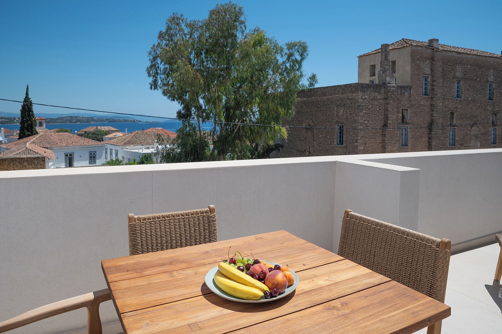 A wooden table with a fruit plate on a balcony overlooking a coastal area with buildings and trees.