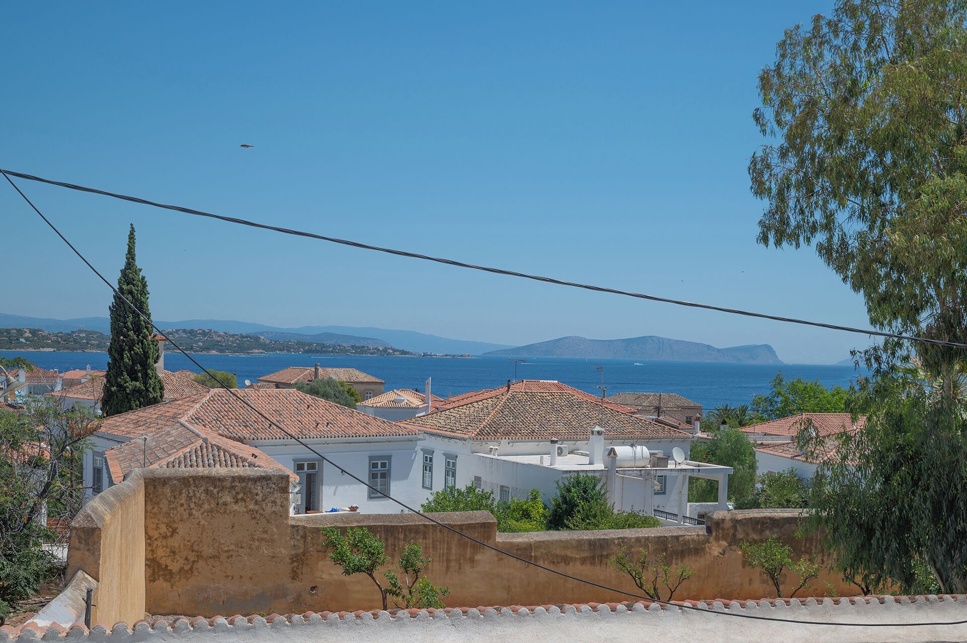 Un quartier résidentiel situé sur la côte, caractérisé par des toits recouverts de tuiles en terre cuite, un cyprès imposant et une vue sur la mer et les montagnes lointaines.