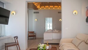 A modern hotel room with a wooden ceiling, a bed with white linens, a beige sofa, a wooden chair, and a small table with a bottle of wine and a glass. - Liberty Suites Spetses  (Spetses)