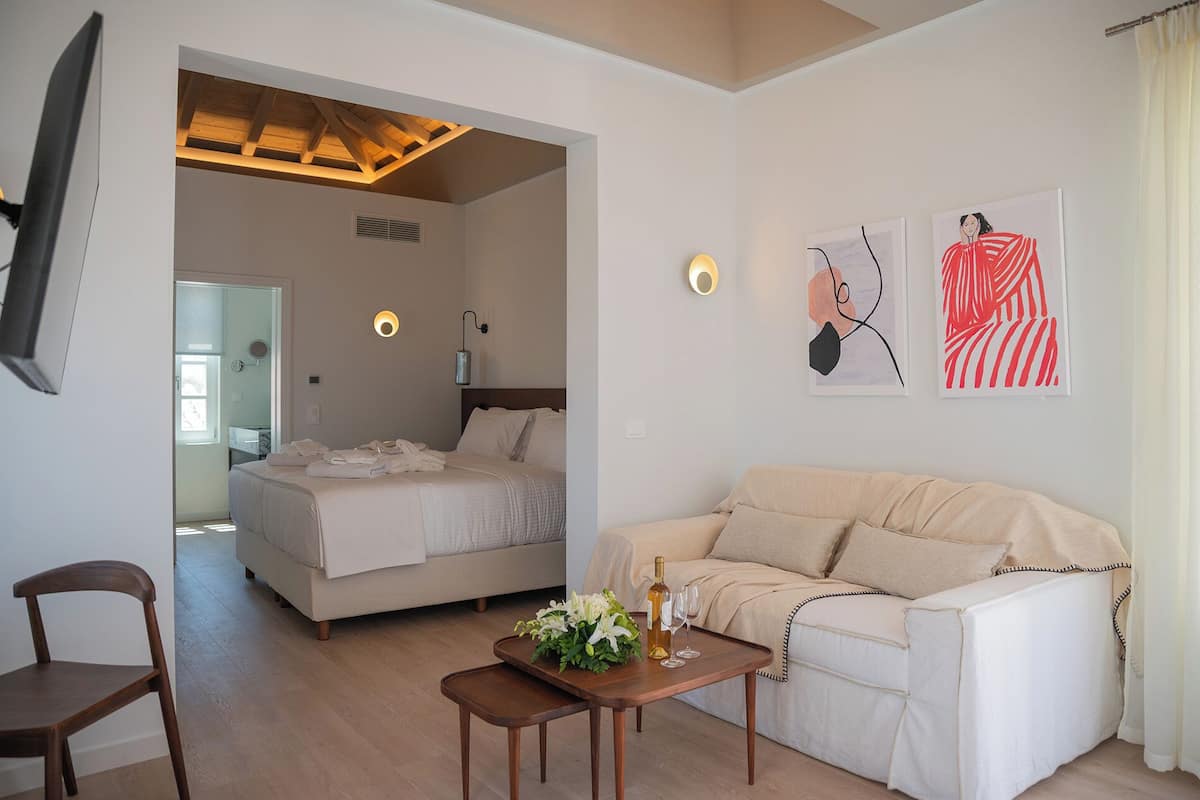 Habitación de hotel con cama, sofá, una mesita de madera con una botella y vasos, y una obra de arte en la pared.