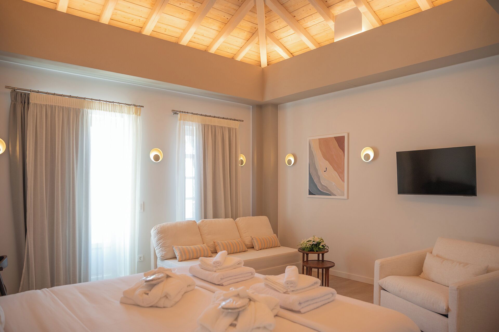 A modern hotel room with a large bed, a sofa, a TV, and a small table with a plant.