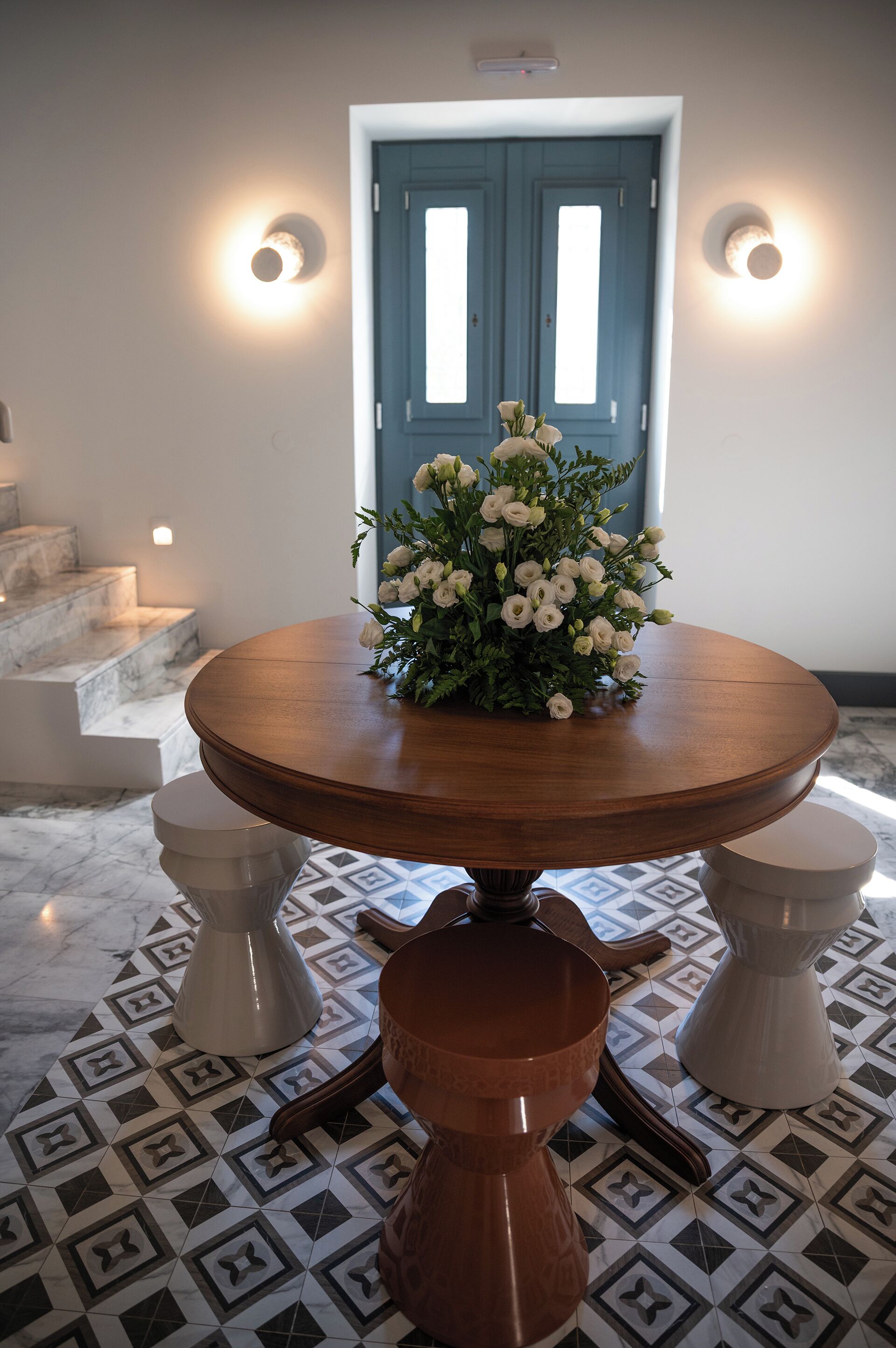 Una mesa de madera redonda con un centro de mesa floral, dos taburetes blancos y un suelo con diseño.