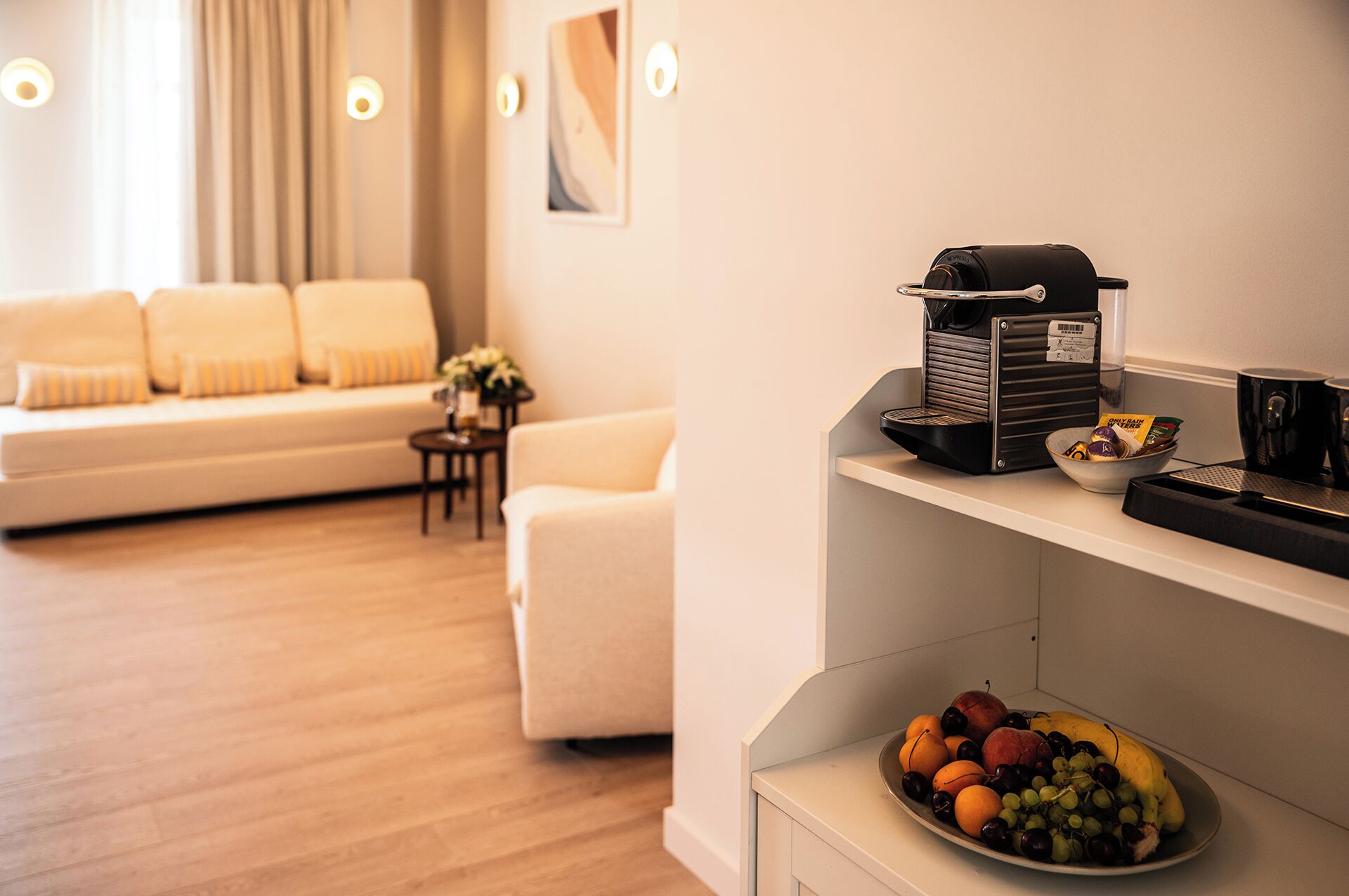 A modern living room with a sofa, a coffee machine, and a bowl of fruit on a shelf.
