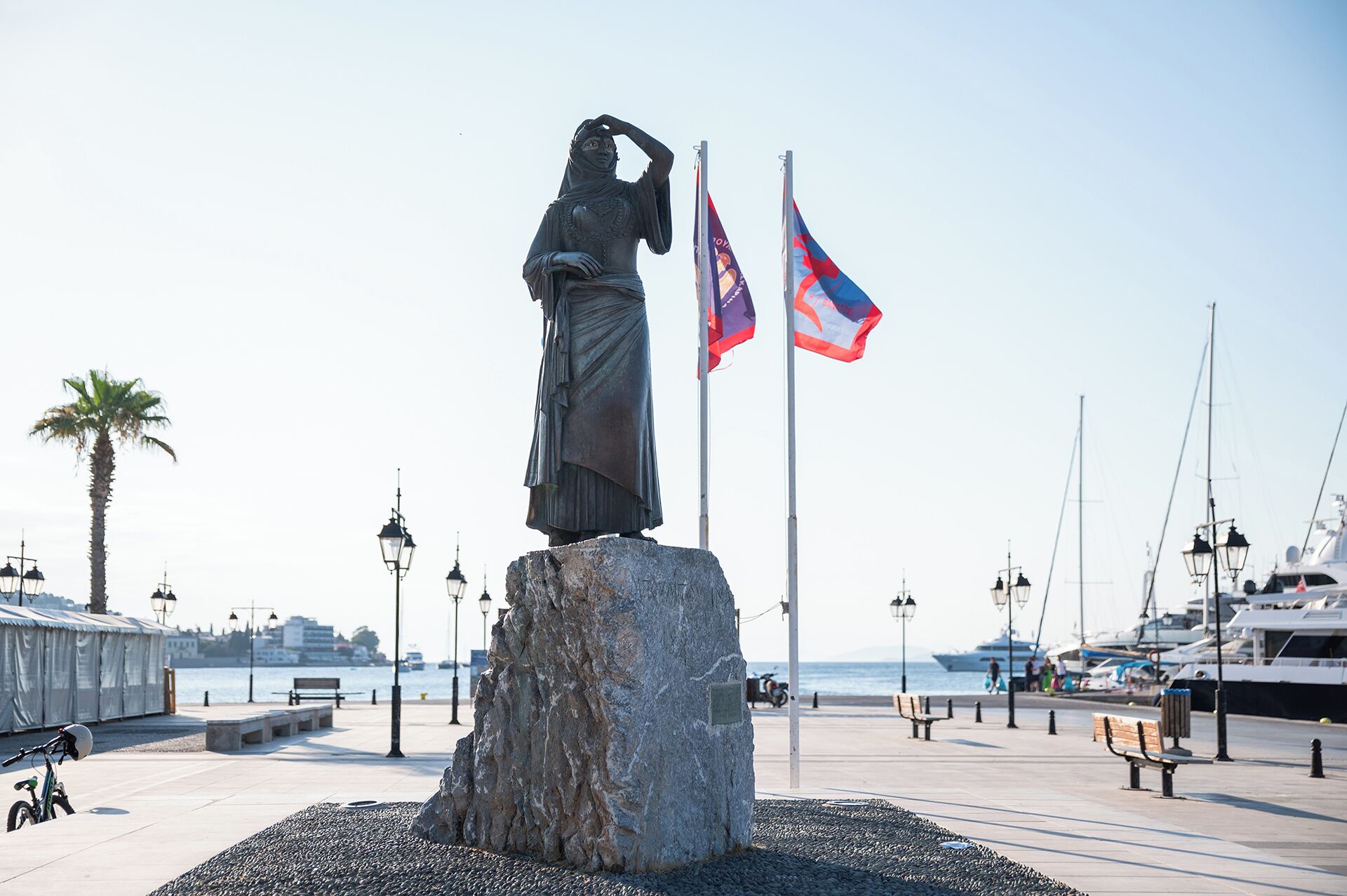 Une statue sur un piédestal en pierre, avec deux drapeaux, un palmier et des bateaux en arrière-plan.