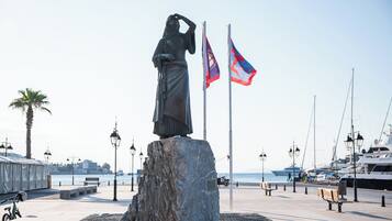 Une statue sur un piédestal en pierre, avec deux drapeaux, un palmier et des bateaux en arrière-plan.