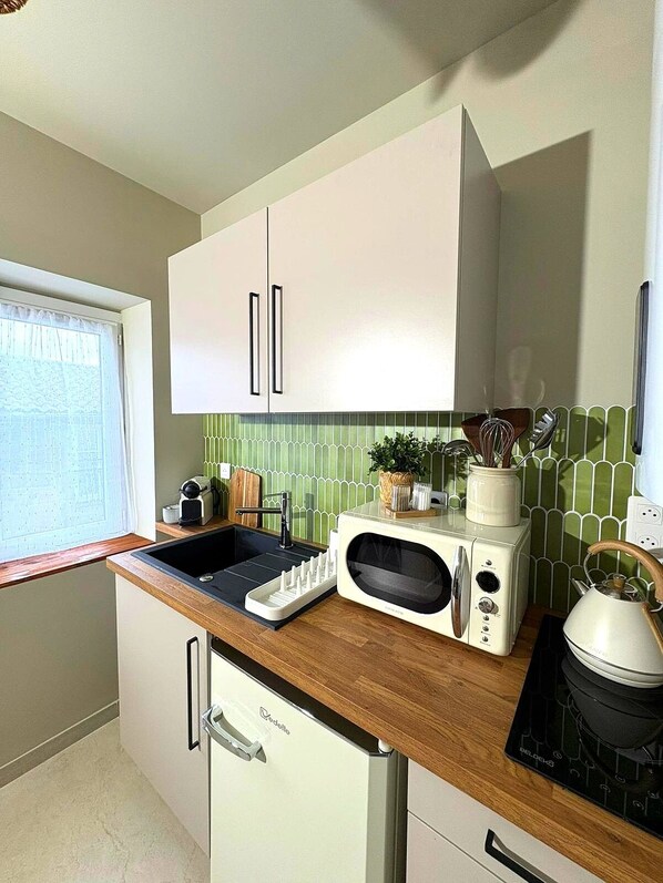 Una cocina con armarios blancos, encimera de madera, microondas integrado, hervidor y lavabo.