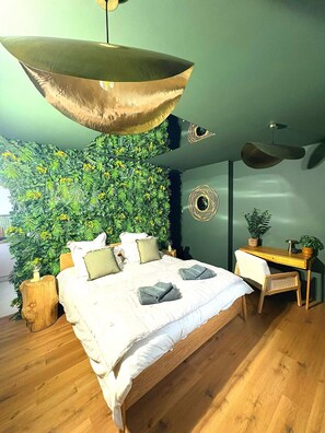 Un dormitorio moderno con una cama grande, un escritorio de madera, una lámpara colgante y una pared verde con follaje.