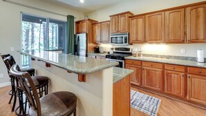 A kitchen with wooden cabinets, a granite countertop, a refrigerator, a microwave, and a stove. There are bar stools and a window with a view of trees. - Tasteful Flagstaff Townhome w/ 2 Furnished Decks (Flagstaff)