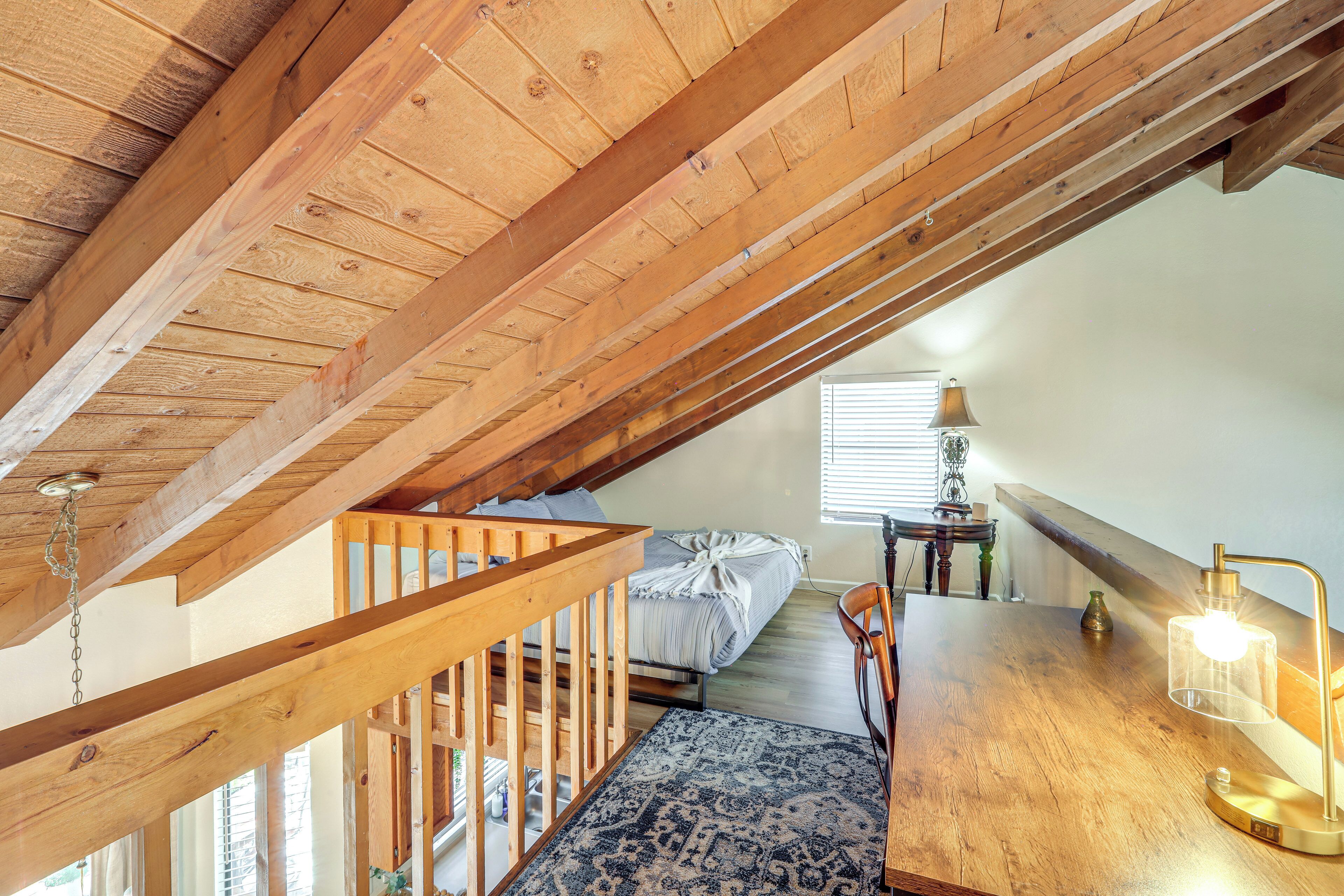 A loft bedroom with exposed wooden beams, a bed, a desk with a lamp, and a view of the outside.
