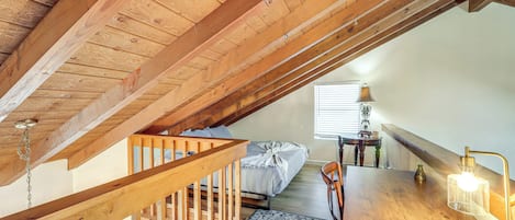 A loft bedroom with exposed wooden beams, a bed, a desk with a lamp, and a view of the outside.