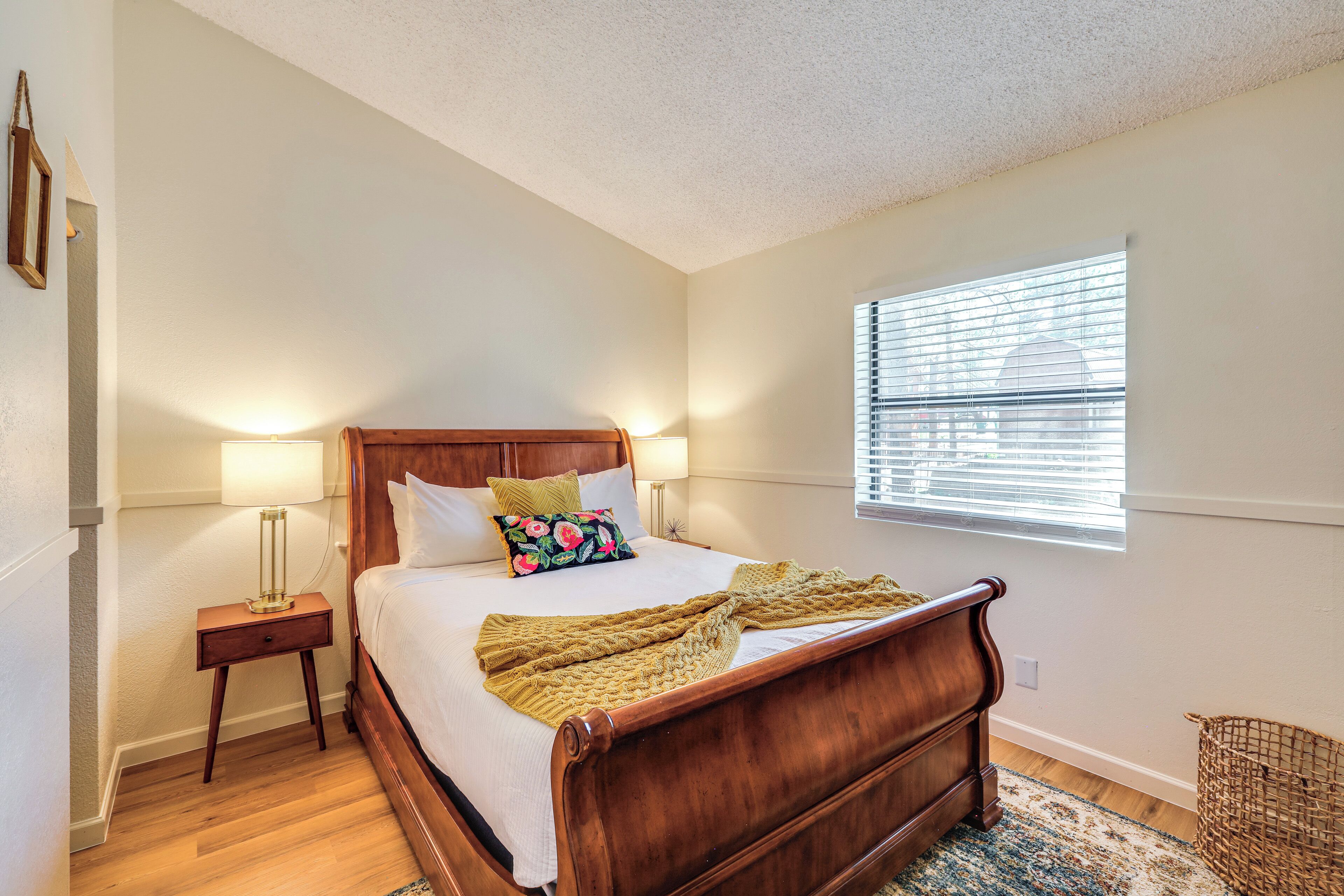 A bedroom with a wooden bed, a nightstand, a lamp, a window with blinds, and a woven basket.