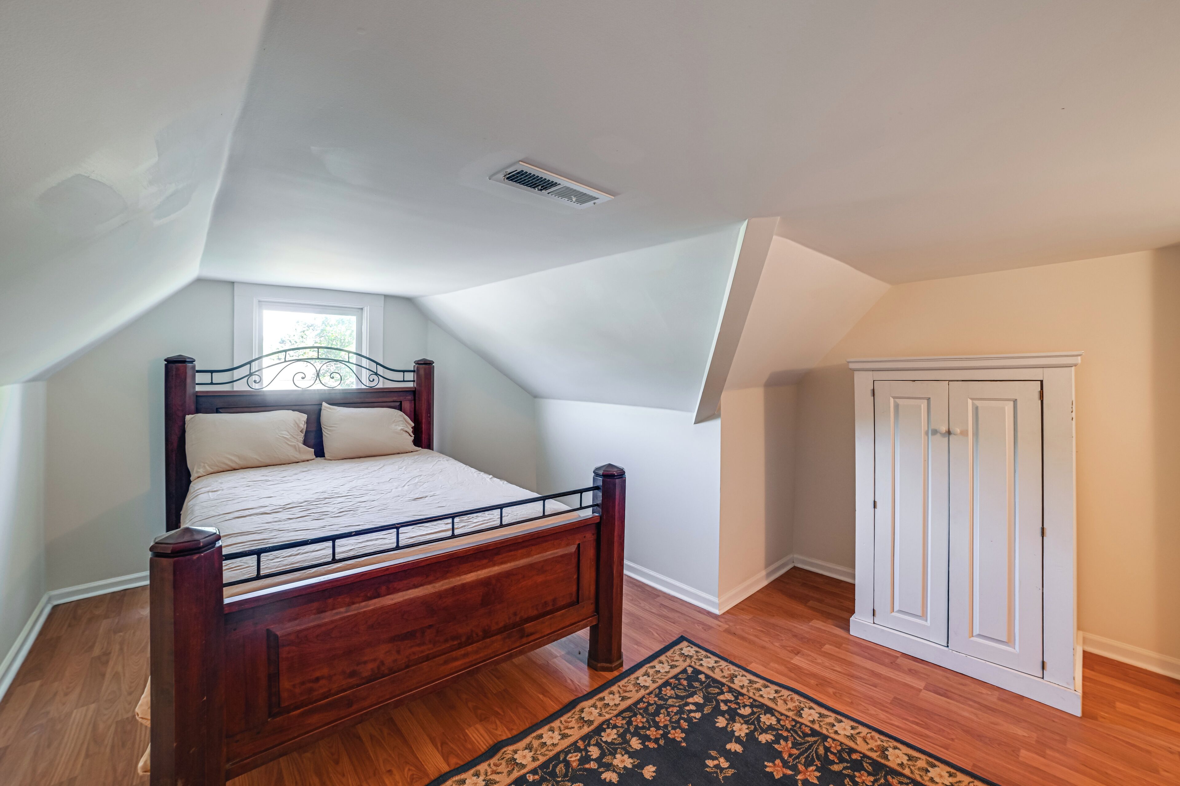 Une chambre à coucher avec un lit en bois, une fenêtre, une porte et une grille d’aération au plafond.
