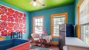 Une pièce aux murs de couleur turquoise, ornée d’une grande fresque florale, avec un ventilateur de plafond, un réfrigérateur, une table avec des chaises et une grande poubelle rouge.