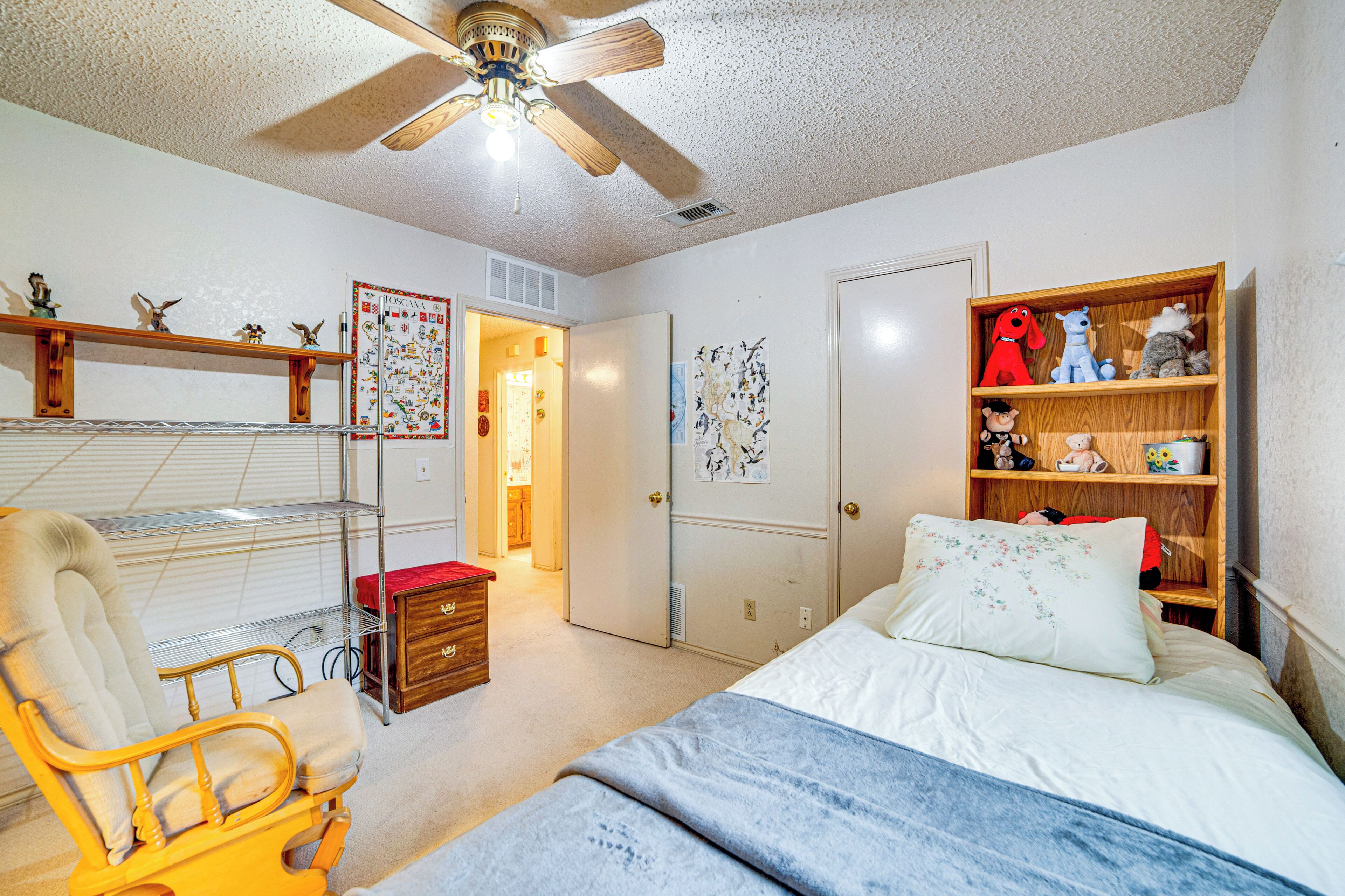 A bedroom with a bed, a chair, a wooden shelf with toys, a ceiling fan, and a door leading to another room.