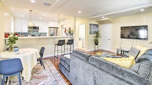A modern living room with a grey sofa, a dining area with a table and chairs, a kitchen with white cabinets, and a flat-screen TV mounted on the wall. - Sunny Oakland Retreat w/ Patio & Cute Garden! (Oakland)