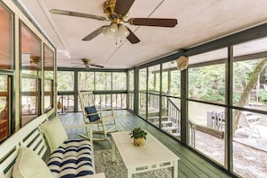 A covered porch with a ceiling fan, a cushioned bench, a table with a plant, and large windows offering a view of the outdoors. - Cartecay River Retreat w/ Yard & Screened Porch! (Ellijay)