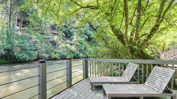 A wooden deck with two lounge chairs overlooking a river, surrounded by lush greenery and a wooden bridge. - Cartecay River Retreat w/ Yard & Screened Porch! (Ellijay)
