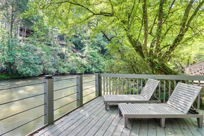 A wooden deck with two lounge chairs overlooking a river, surrounded by lush greenery and a wooden bridge. - Cartecay River Retreat w/ Yard & Screened Porch! (Ellijay)