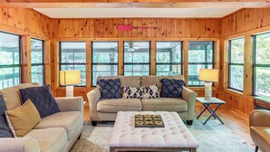 A cozy room with a beige sofa, a matching ottoman, and a small side table with a lamp. Large windows offer a view of the outdoors. - Cartecay River Retreat w/ Yard & Screened Porch! (Ellijay)