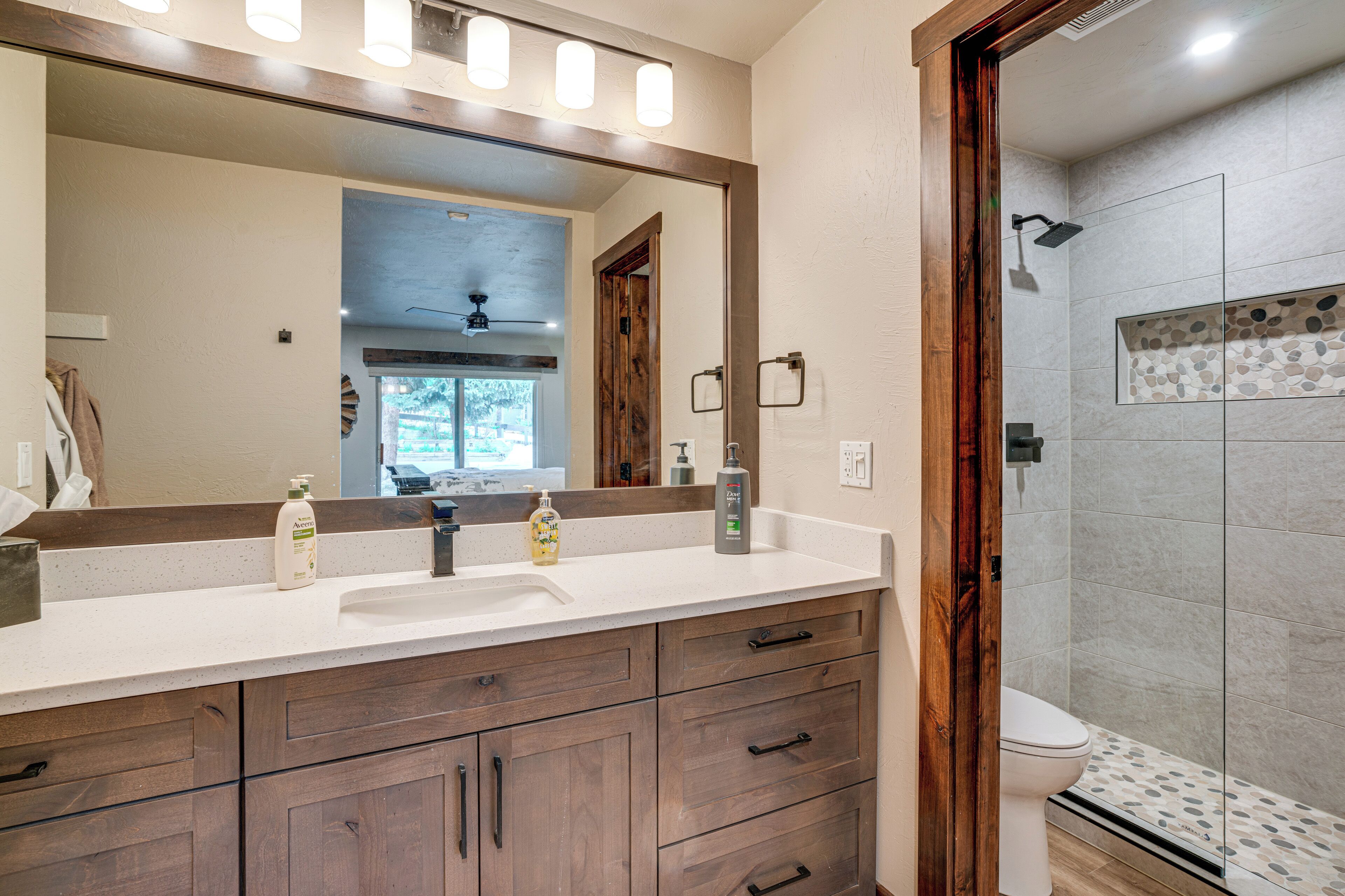 A bathroom with a large mirror, wooden cabinets, a white sink, and a shower area with patterned tiles.