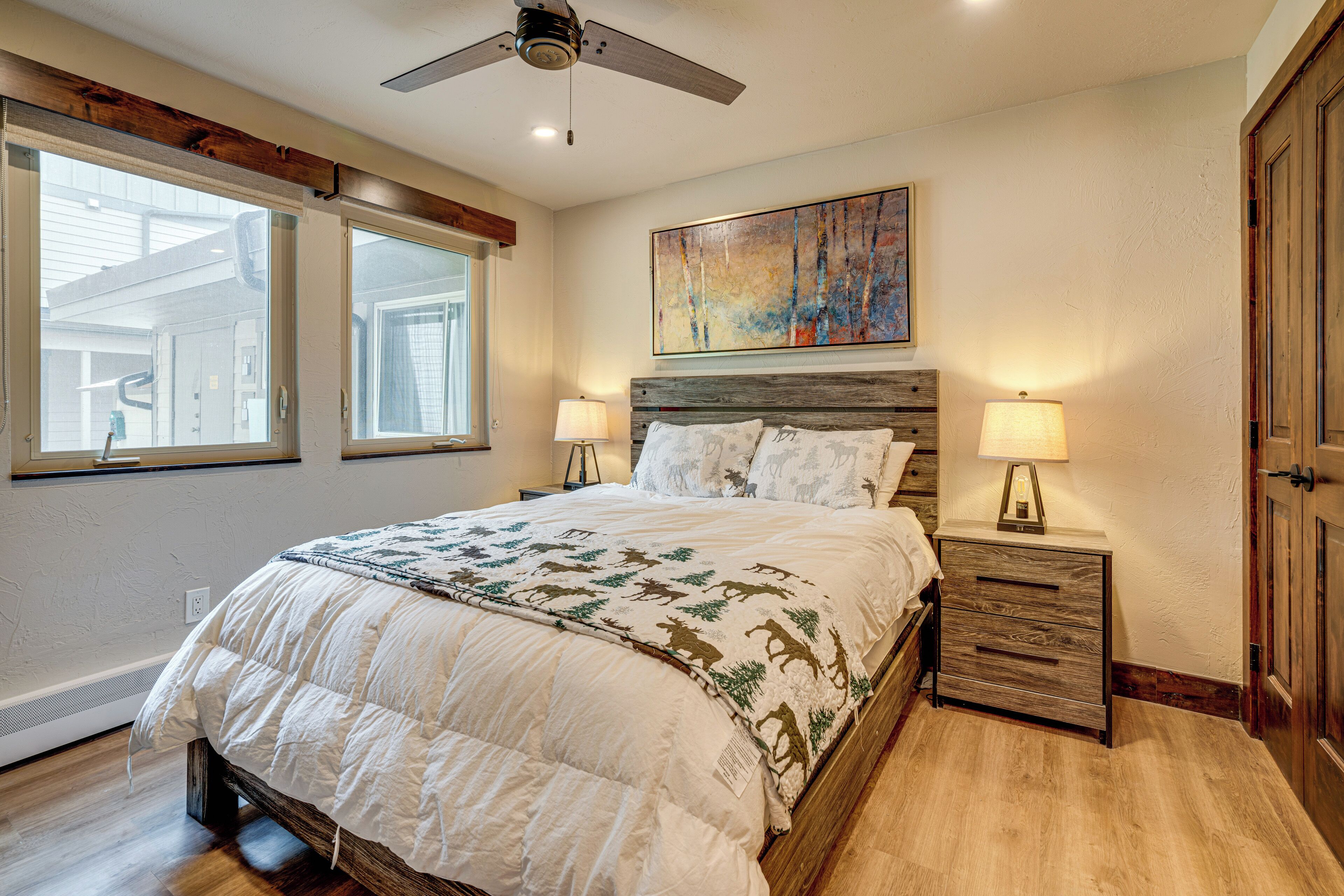 A bedroom with a large bed, two bedside tables with lamps, a ceiling fan, and a painting on the wall.