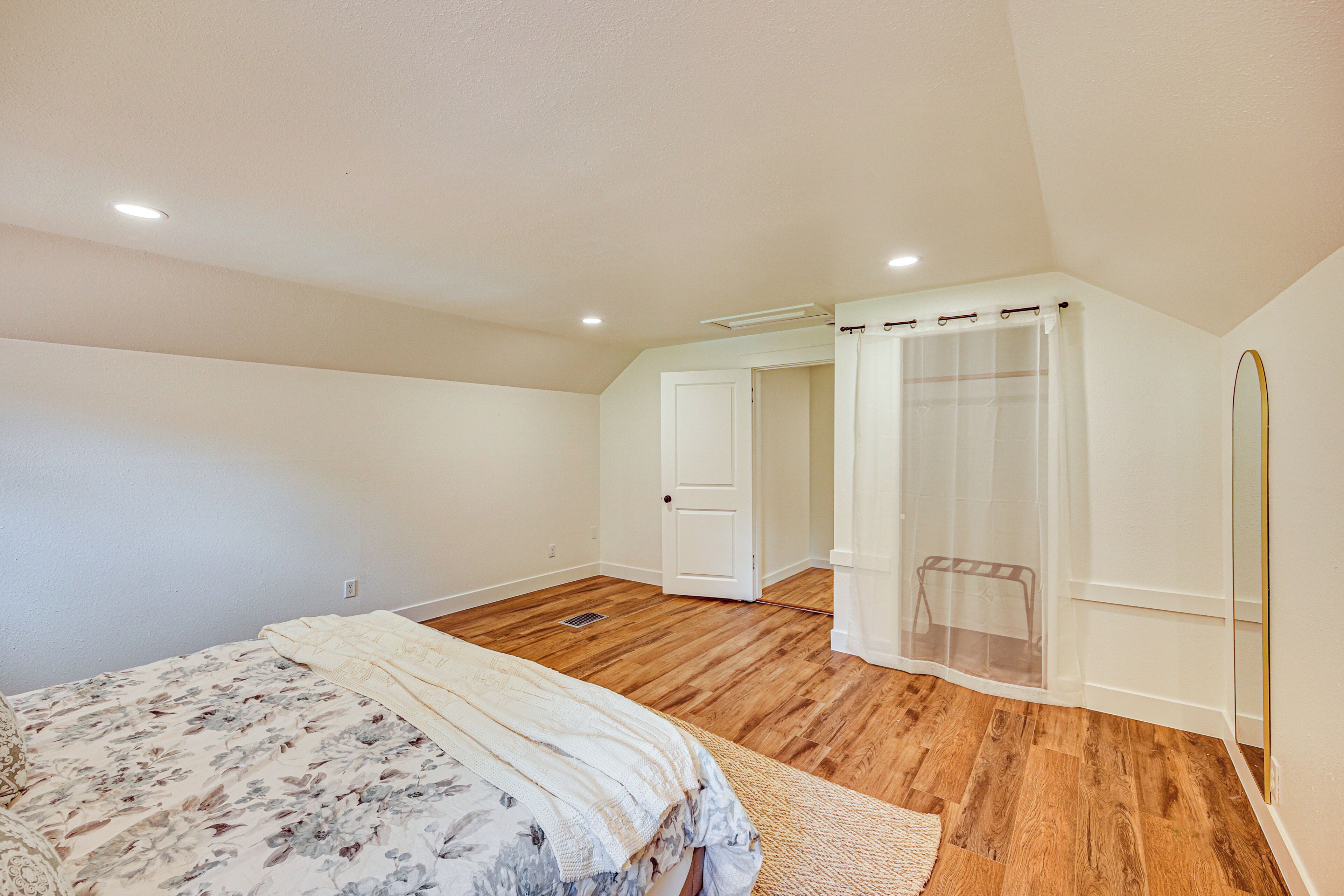 A bedroom with a wooden floor, a bed with a floral bedspread, a white door, and a shower area with a curtain.