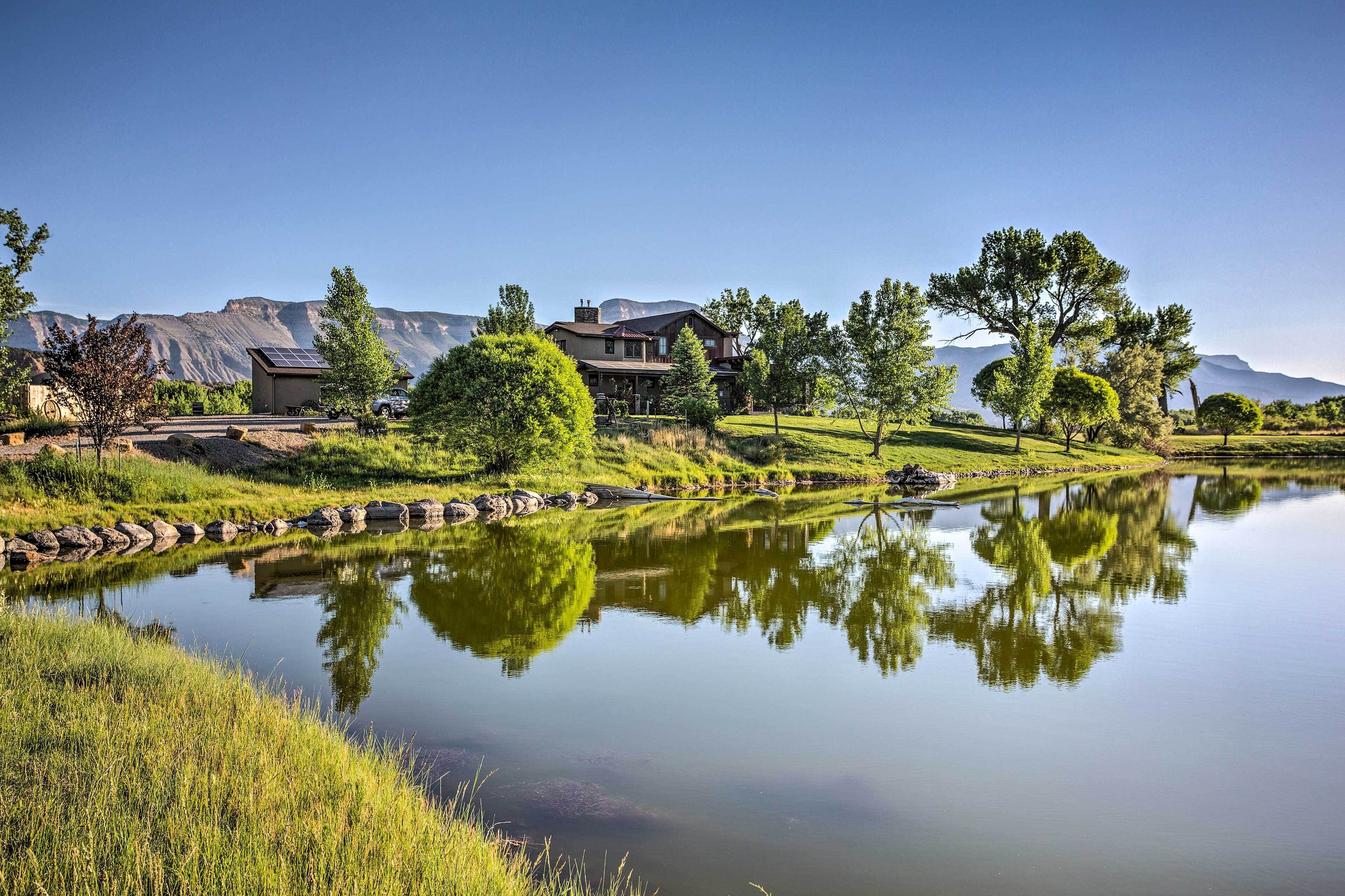 A large house with a pond in front, surrounded by trees and mountains.