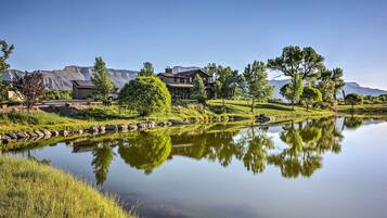 A large house with a pond in front, surrounded by trees and mountains.