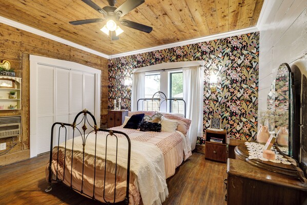 A bedroom with a wooden ceiling, a bed with a metal frame, a window with curtains, and a floral wallpaper.