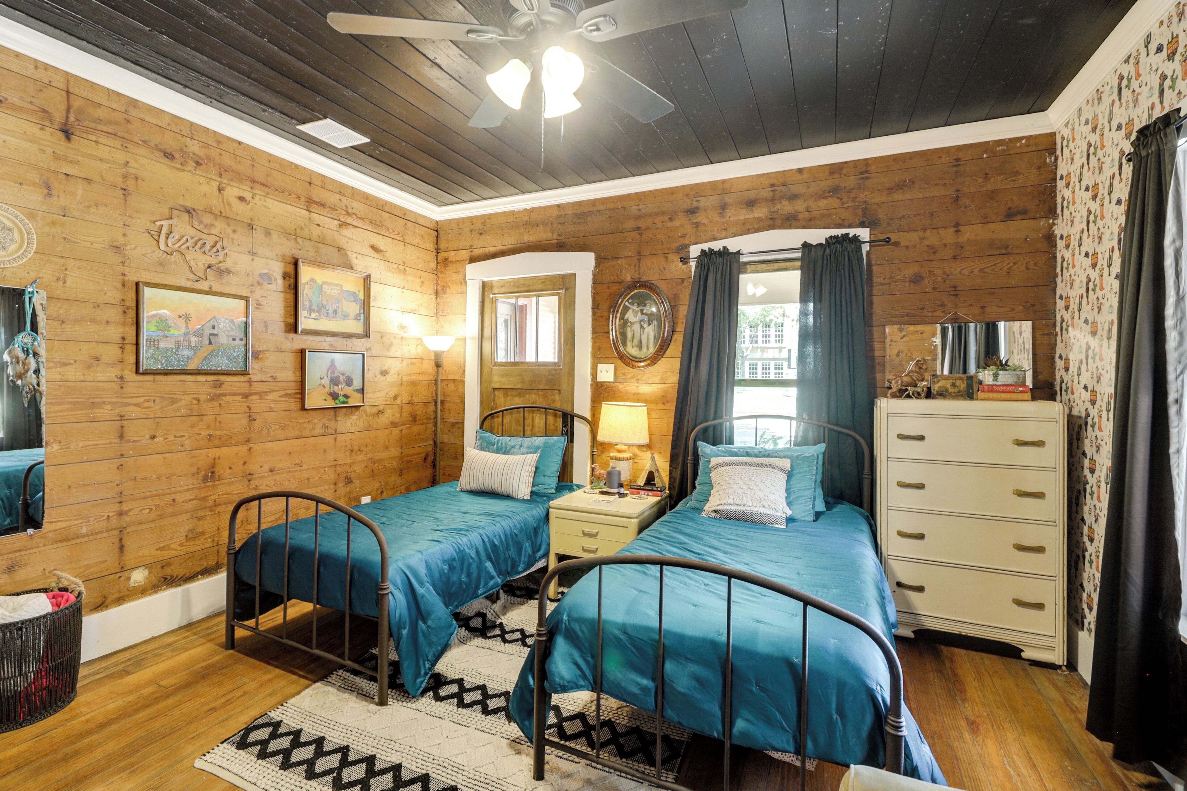 Una habitación acogedora con paredes de madera, dos camas con ropa de cama azul a juego, una mesita de noche con lámpara, un aparador y un ventilador de techo.