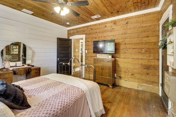A bedroom with a wooden ceiling, a bed with a pink and white bedspread, a dresser, a television on the wall, and a door leading to another room. - 1925 Craftsman Bungalow - 1 Mi to Dtwn Waxahachie! (Waxahachie)