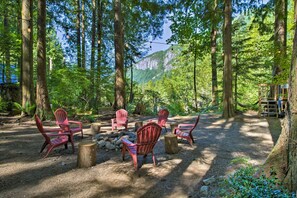 Waldlandschaft mit roten Adirondack-Stühlen, die um eine zentrale Feuerstelle angeordnet sind, umgeben von Bäumen und einem Berg im Hintergrund.