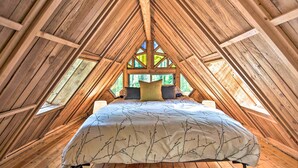 Une chambre mansardée chaleureuse, avec un plafond en pente en bois, un lit recouvert d’une couvre-lit à motifs de branches et une vue sur la verdure grâce à une fenêtre triangulaire.