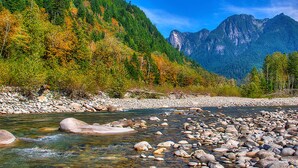 Une rivière aux eaux claires, bordée de rochers, entourée d’arbres verdoyants et avec des montagnes en arrière-plan.