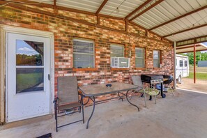 Een overdekt terras met een bakstenen muur, een tafel met stoelen, een barbecue en een geparkeerde camper.