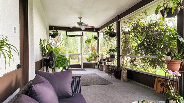 Une terrasse couverte avec un ventilateur de plafond, un canapé avec des coussins et diverses plantes en pot.