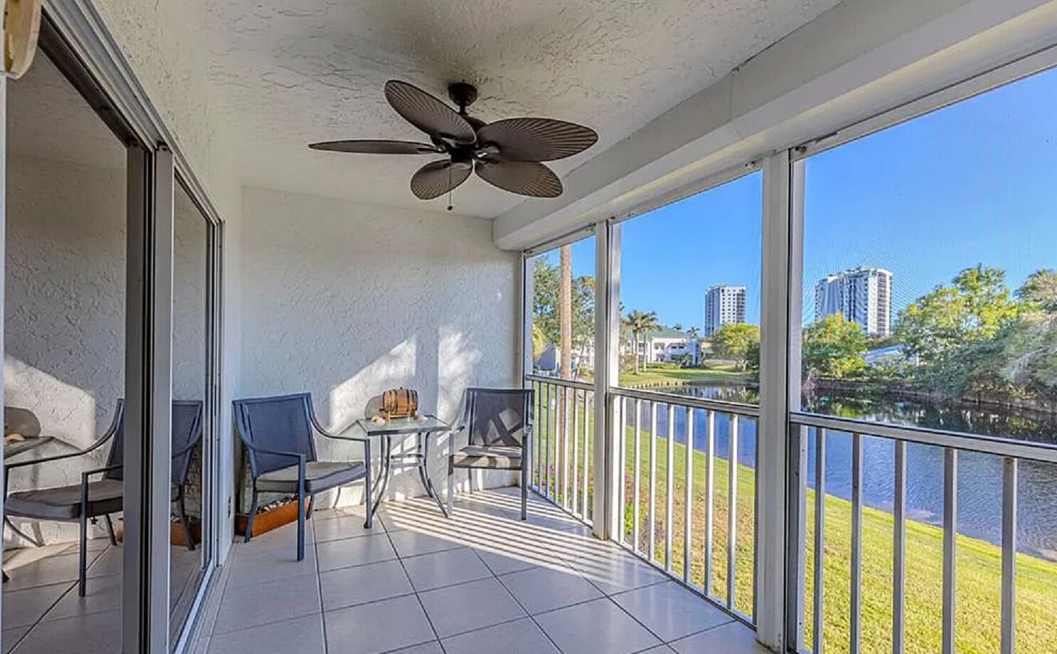 A balcony with a ceiling fan, two chairs, a small table, and a view of a lake and buildings.