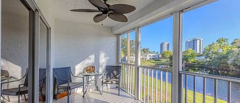 A balcony with a ceiling fan, two chairs, a small table, and a view of a lake and buildings.