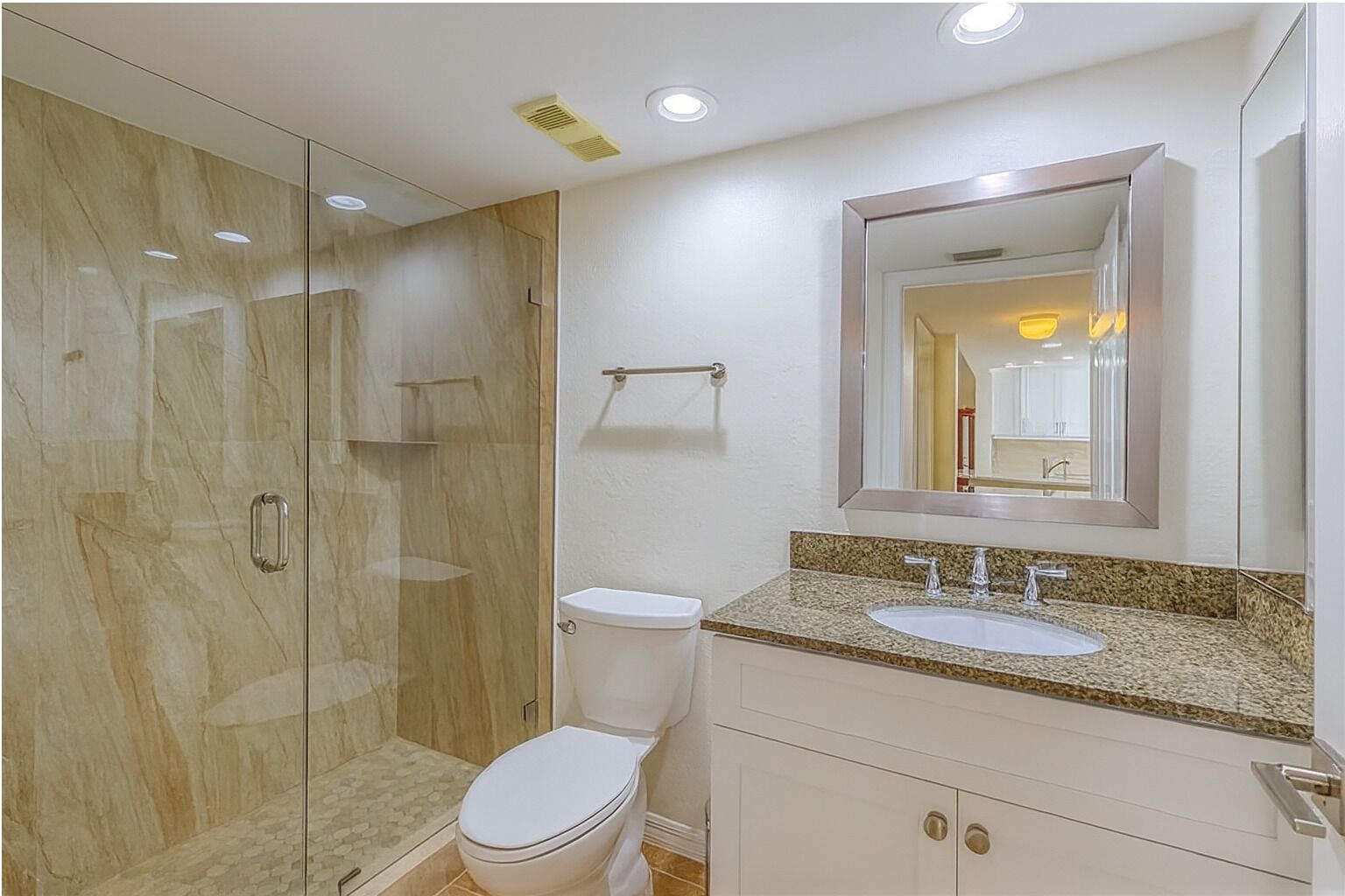 Une salle de bain moderne avec une douche à parois de verre, des toilettes blanches, un lavabo sur un plan de travail en granit et un grand miroir.