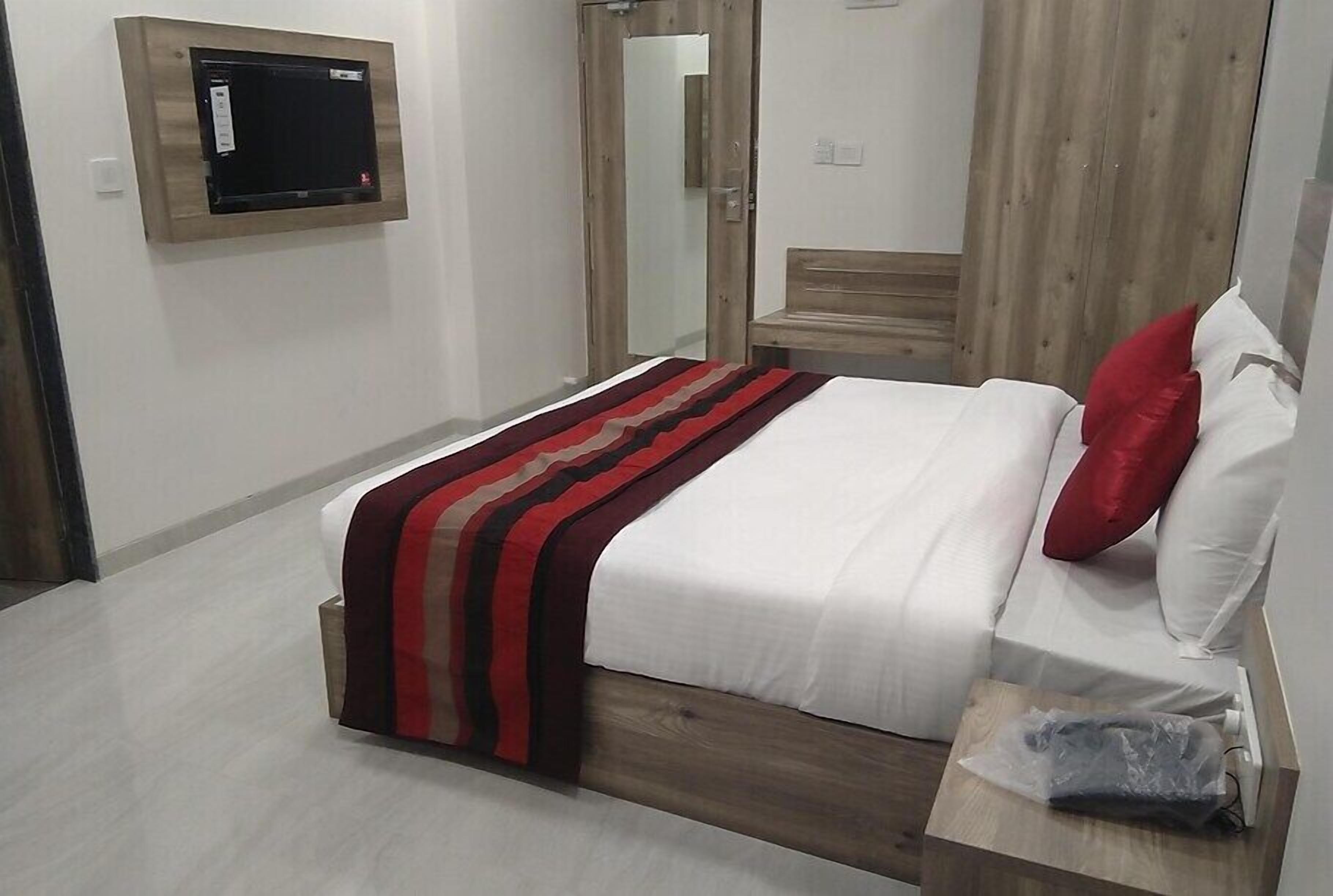 Quarto com cama, cabeceira de madeira, mesa de cabeceira com saco plástico, televisão na parede e espelho.
