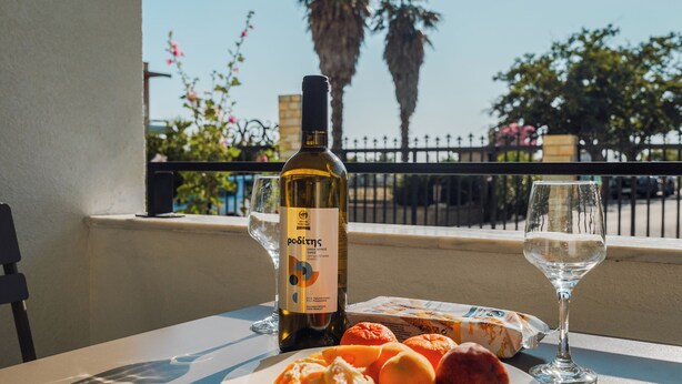 Une table avec une bouteille de vin, un verre et quelques oranges, sur un balcon d’où l’on aperçoit des palmiers et une clôture.