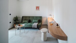 Un salon moderne avec un canapé vert, une petite table blanche, un tabouret rond blanc et un buffet en bois surmonté d’une lampe.