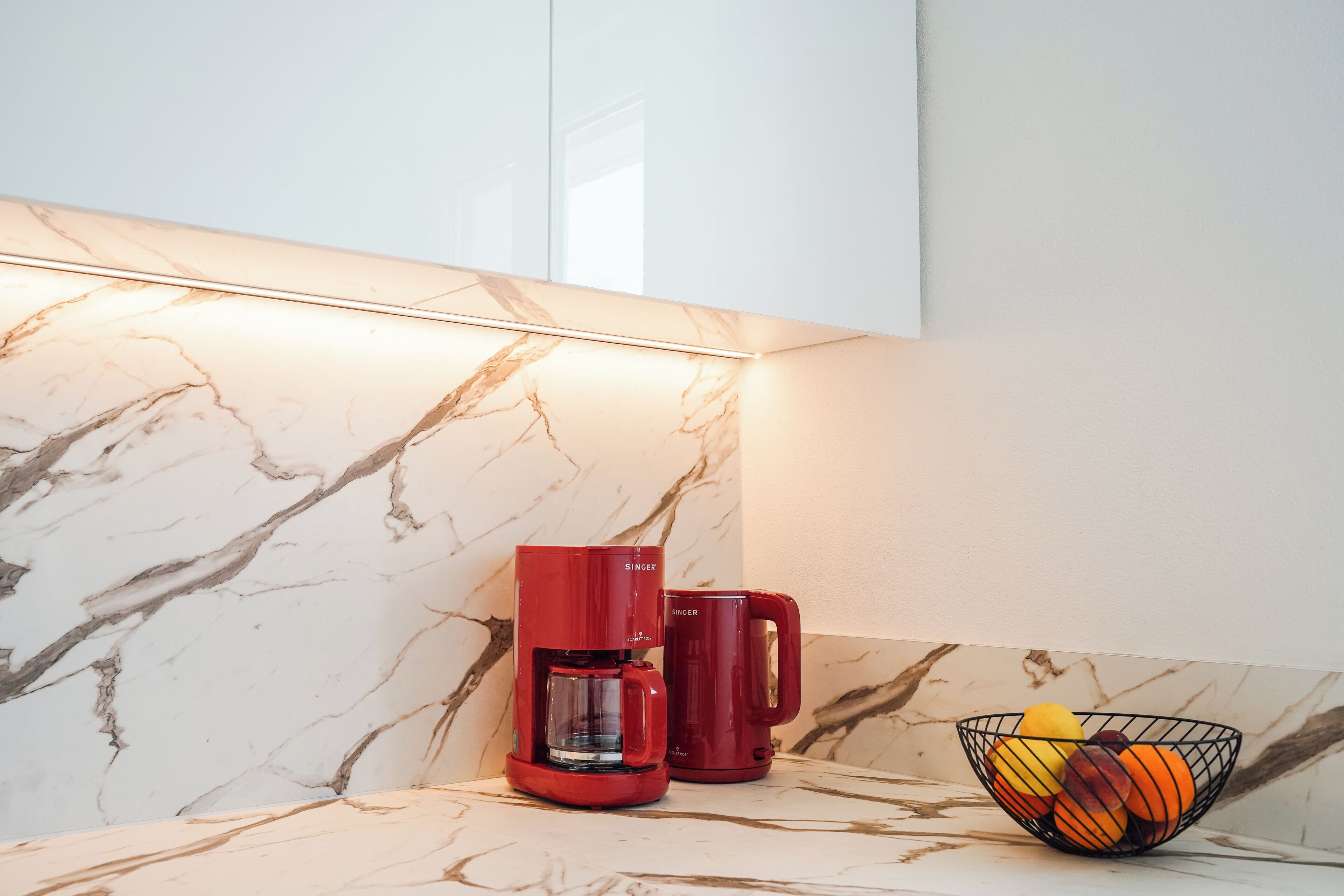 A kitchen with a marble backsplash, a red coffee maker, and a fruit bowl.