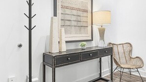 Ein Zimmer mit einer schwarzen Konsole, einem gerahmten Kunstwerk, einer Lampe, einem Sessel und einem Kleiderhaken.
