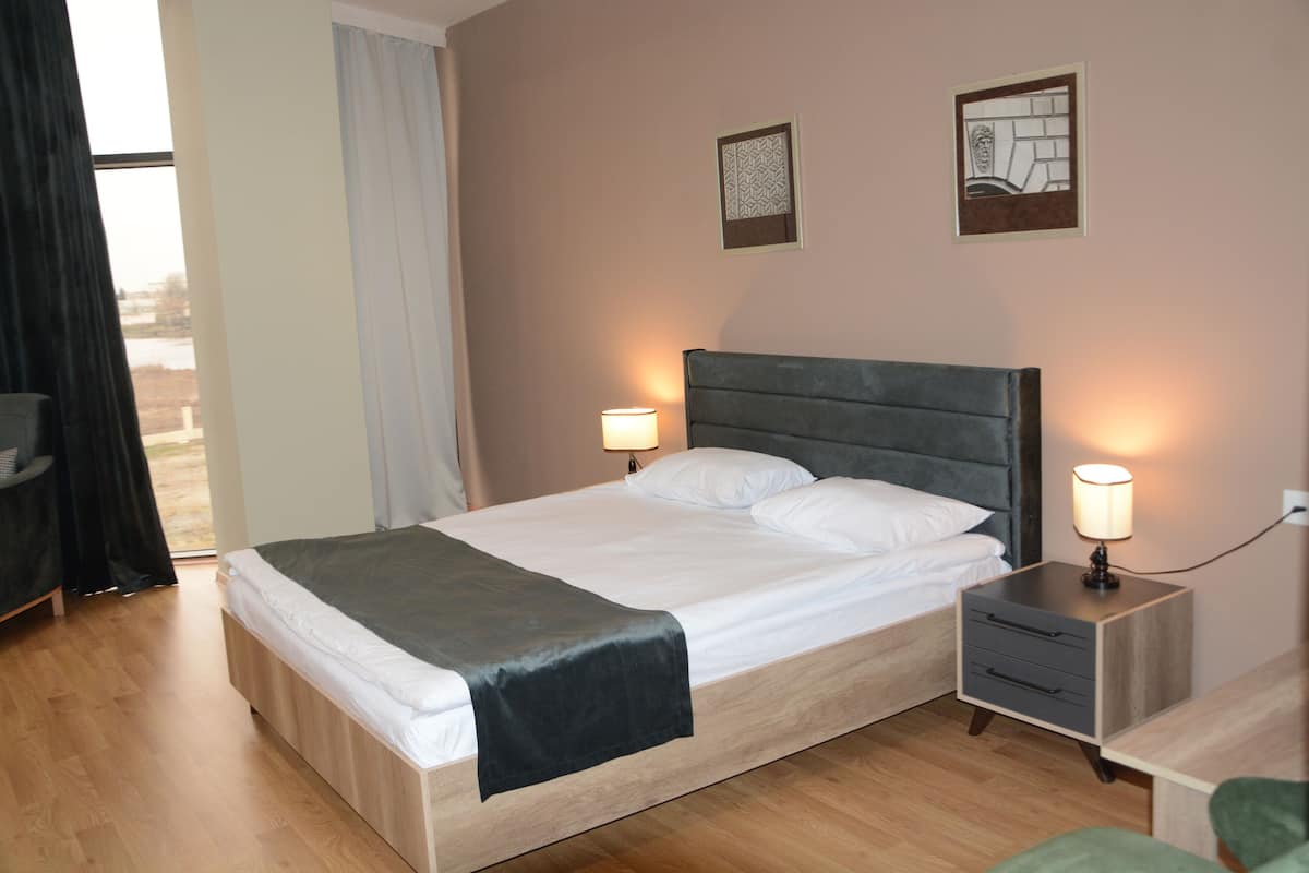 Uma cama de casal com cabeceira cinza, duas mesas de cabeceira com abajures e piso de madeira.