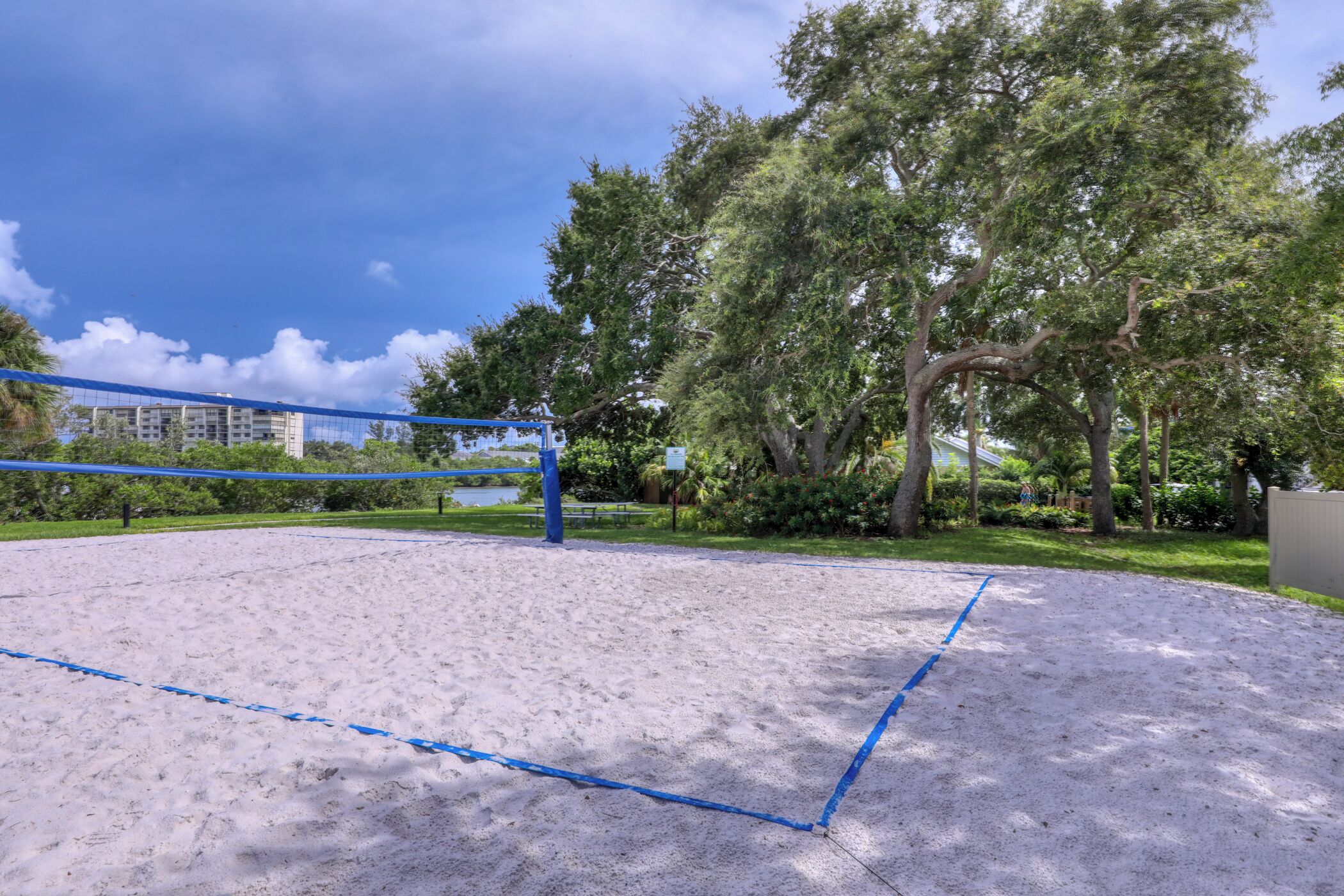 A sandy outdoor volleyball court with a blue net, surrounded by trees and greenery.