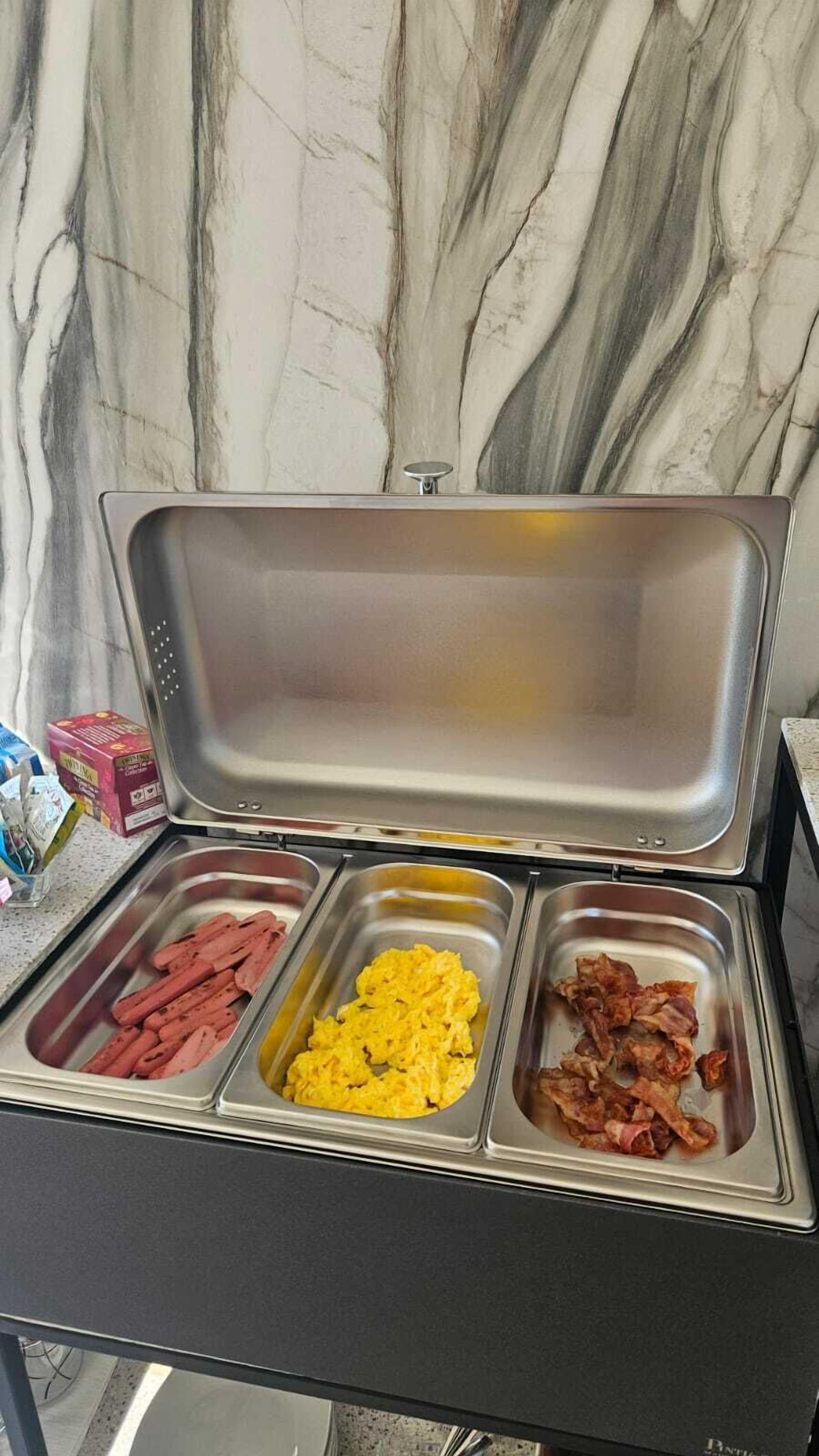 Un bufé con tres fuentes de comida que incluyen salchichas, huevos revueltos y tocino.