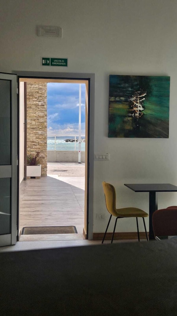 A room with a view of the sea, a painting on the wall, and a sign indicating an emergency exit.