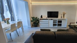 A modern living room with a sofa, a TV on a white cabinet, a dining area with chairs, and a potted plant.