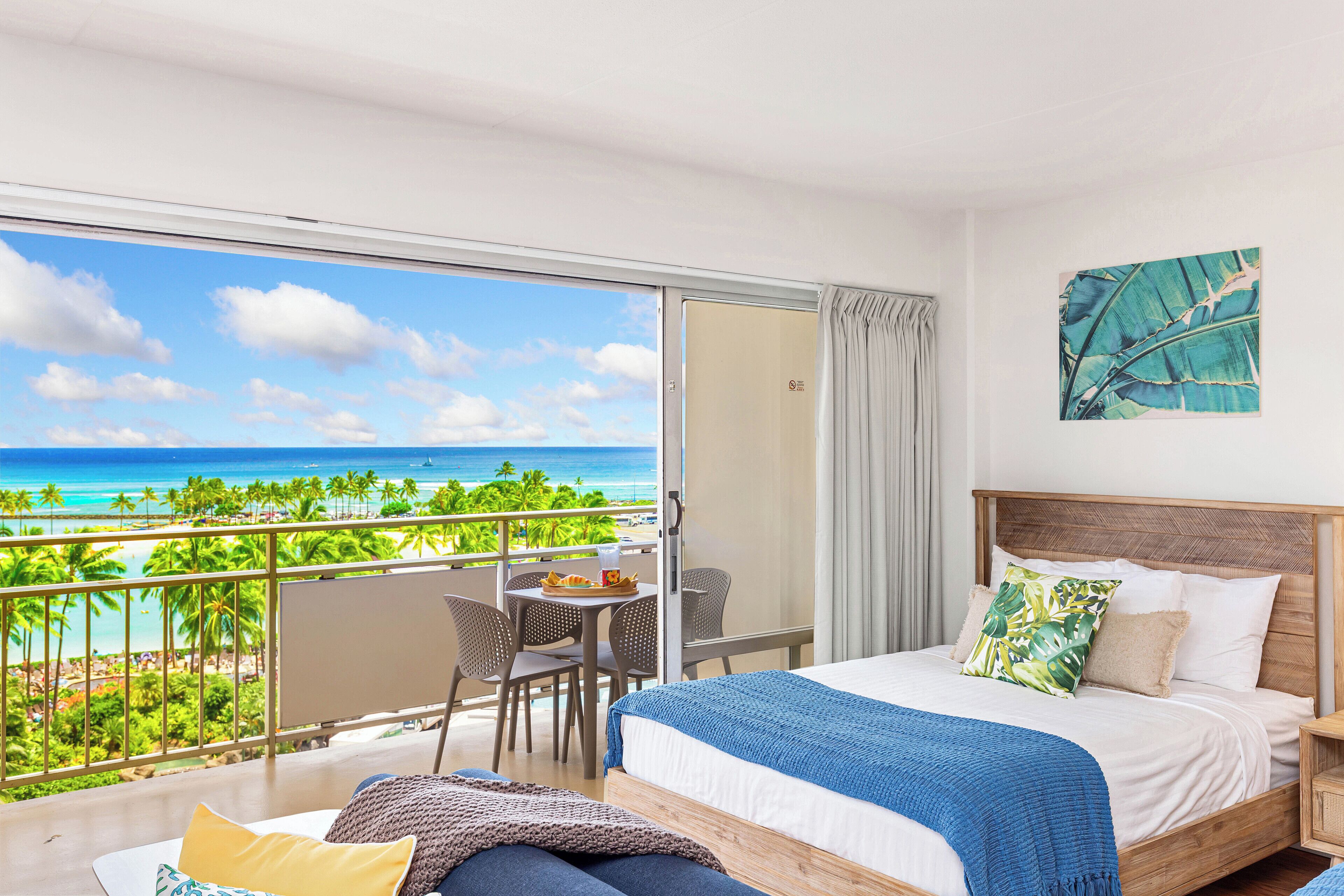 A hotel room with a balcony overlooking a beach, a bed with a wooden headboard, and a table with chairs.
